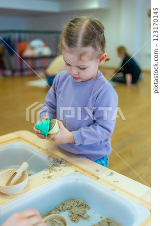 Little girl playing with kinetic sand and wooden toys. Sensory development and experiences, themed activities with children, fine motor skills development Little girl playing with kinetic sand and wooden toys. Sensory development and experiences, themed activities with children, fine motor skills development 123170145