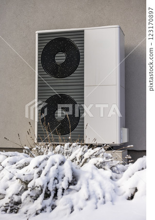 Close-up of a dual-fan outdoor heat pump mounted on a gray wall, surrounded by snow-covered bushes, illustrating modern energy efficiency. 123170897