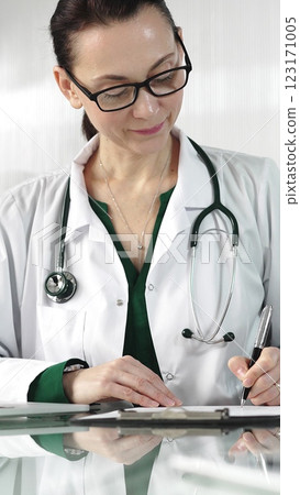 Female doctor in a green blouse under a medical coat is filling up medication history records on a glass desk in the clinic, vertical portrait view. Medicine concept Female doctor in a green blouse under a medical coat is filling up medication history records on a glass desk in the clinic, vertical portrait view. Medicine concept 123171005