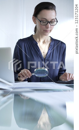 Woman accountant examining financial documents with magnifying glass at glass office desk, conducting thorough analysis of business transactions. Audit and taxes in business Woman accountant examining financial documents with magnifying glass at glass office desk, conducting thorough analysis of business transactions. Audit and taxes in business 123171204