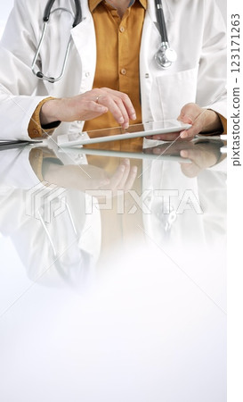 Unknown male doctor wearing a stethoscope, yellow shirt under a lab coat, is using digital tablet in modern healthcare setting, interacting with patient data, vertical portrait. Medicine concept 123171263