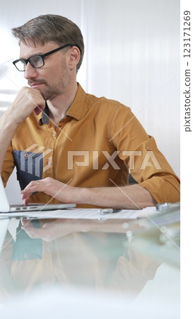 Focused businessman in yellow casual shirt is using laptop and thinking with hand on chin, working on a project in a modern office, vertical portrait. Business people concept Focused businessman in yellow casual shirt is using laptop and thinking with hand on chin, working on a project in a modern office, vertical portrait. Business people concept 123171269