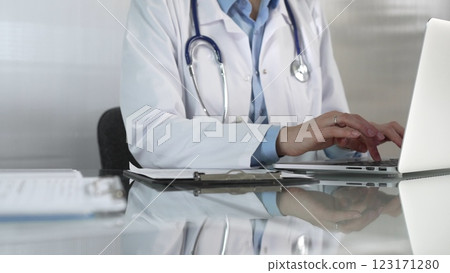 Doctor woman, wearing a blue blouse under a medical coat, is using technology in healthcare, close up of hands typing on laptop with stethoscope, glasses, and clipboard on desk. Medicine concept Doctor woman, wearing a blue blouse under a medical coat, is using technology in healthcare, close up of hands typing on laptop with stethoscope, glasses, and clipboard on desk. Medicine concept 123171280