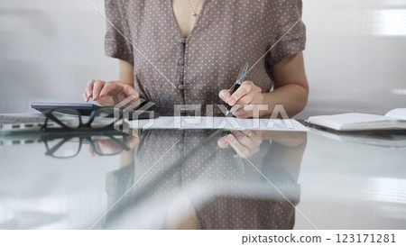 Businesswoman accountant, wearing brawn dotted blouse, is using a calculator to crunch numbers and jotting down meticulous notes, at sleek glass desk in a contemporary office. Taxes, audit in business 123171281