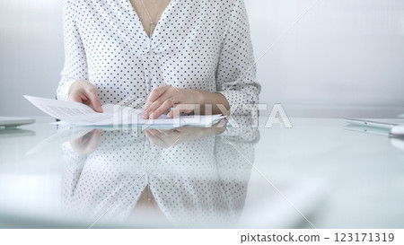 Businesswoman in white dotted blouse is going through paperwork while holding documents at a glass desk in a well lit, contemporary office setting. Business people concept 123171319