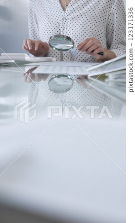 Businesswoman in white dotted blouse is carefully reviewing financial reports with magnifying glass and calculator, conducting audit and analyzing data, vertical view Taxes, audit in business 123171336
