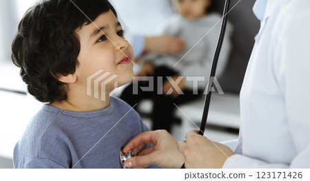 Doctor-woman examining a child patient by stethoscope. Cute arab boy and his brother at physician appointment. Medicine concept Doctor-woman examining a child patient by stethoscope. Cute arab boy and his brother at physician appointment. Medicine concept 123171426