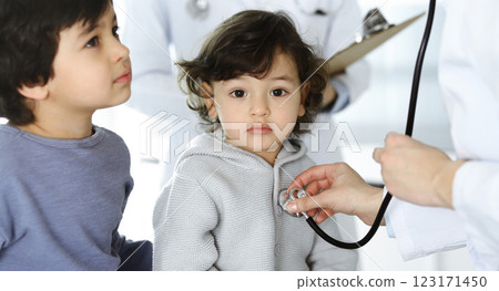 Woman-doctor examining a child patient by stethoscope. Cute arab toddler and his brother at physician appointment. Medicine concept Woman-doctor examining a child patient by stethoscope. Cute arab toddler and his brother at physician appointment. Medicine concept 123171450