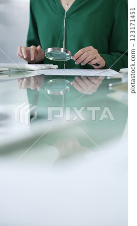 Businesswoman in green blouse carefully reviewing financial reports with magnifying glass and calculator, conducting audit and analyzing data in office. Taxes, audit in business 123171451