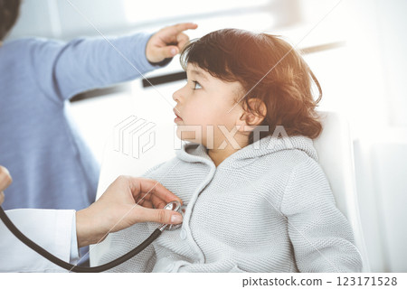 Doctor-woman examining a child patient by stethoscope in sunny clinik. Cute arab boy and his brother at physician appointment 123171528