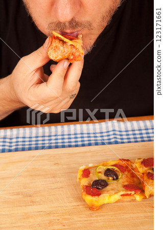 Bearded caucasian white mature male in black t-shirt eating delicious pizza slices on bamboo cutting board on table 123171661