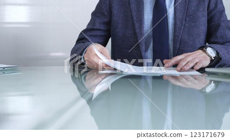 Businessman wearing blue jacket is reviewing important paperwork while holding documents at a sleek glass desk in a modern office environment. Business people or audit concept Businessman wearing blue jacket is reviewing important paperwork while holding documents at a sleek glass desk in a modern office environment. Business people or audit concept 123171679