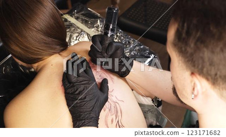 Professional tattoo artist wearing black gloves drawing intricate design on woman's back while working in sleek, well lit tattoo studio with modern equipment 123171682