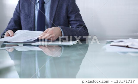 Businessman wearing blue jacket is reviewing important paperwork while holding documents at a sleek glass desk in a modern office environment. Business people or audit concept Businessman wearing blue jacket is reviewing important paperwork while holding documents at a sleek glass desk in a modern office environment. Business people or audit concept 123171737