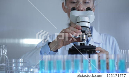 A female scientist, wearing a lab coat and safety glasses, is adjusting a binocular microscope surrounded by test tubes in laboratory setting. Medicine and science A female scientist, wearing a lab coat and safety glasses, is adjusting a binocular microscope surrounded by test tubes in laboratory setting. Medicine and science 123171757