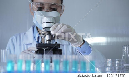 A scientist woman, wearing a lab coat, mask, white gloves and safety glasses, is adjusting a microscope surrounded by test tubes in laboratory A scientist woman, wearing a lab coat, mask, white gloves and safety glasses, is adjusting a microscope surrounded by test tubes in laboratory 123171761