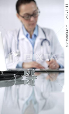 Stethoscope and medication history records form are lying on a glass desk before a doctor working with laptop in clinical office background, vertical close-up view. Medicine and science 123171811