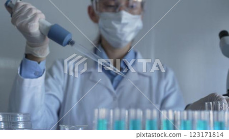 Scientist woman, wearing a lab coat, mask, blue gloves and safety glasses, is using a micropipette to drop a liquid into a test tubes. Medicine and science Scientist woman, wearing a lab coat, mask, blue gloves and safety glasses, is using a micropipette to drop a liquid into a test tubes. Medicine and science 123171814