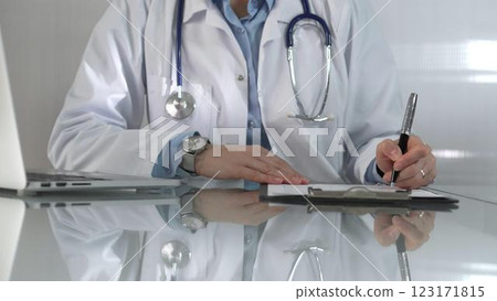 Woman doctor, wearing a blue blouse under a white medical coat, is using a clipboard and taking notes on glass desk in a medical office, close-up. Medicine Woman doctor, wearing a blue blouse under a white medical coat, is using a clipboard and taking notes on glass desk in a medical office, close-up. Medicine 123171815