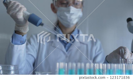 Scientist woman, wearing a lab coat, mask, blue gloves and safety glasses, is using a micropipette to drop a liquid into a test tubes. Medicine and science 123171836