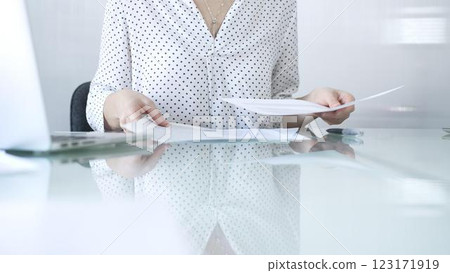Businesswoman wearing white dotted blouse is going through paperwork while holding documents at a glass desk in a modern office setting. Business people concept 123171919