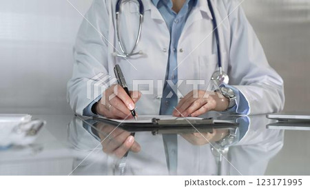 Woman doctor, wearing a blue blouse under a white medical coat, is using a clipboard and taking notes on glass desk in a medical office, close-up. Medicine Woman doctor, wearing a blue blouse under a white medical coat, is using a clipboard and taking notes on glass desk in a medical office, close-up. Medicine 123171995