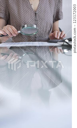 Businesswoman accountant in brawn dotted blouse reviewing financial reports with magnifying glass and calculator in office, vertical view. Taxes, audit in business Businesswoman accountant in brawn dotted blouse reviewing financial reports with magnifying glass and calculator in office, vertical view. Taxes, audit in business 123172003
