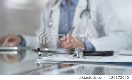 Stethoscope and medication history records form are on a glass table before a male doctor working with laptop in clinical office background. Medicine concept 123172030