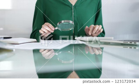 Businesswoman in green blouse carefully reviewing financial reports with magnifying glass and calculator, conducting audit and analyzing data in office. Taxes, audit in business Businesswoman in green blouse carefully reviewing financial reports with magnifying glass and calculator, conducting audit and analyzing data in office. Taxes, audit in business 123172034