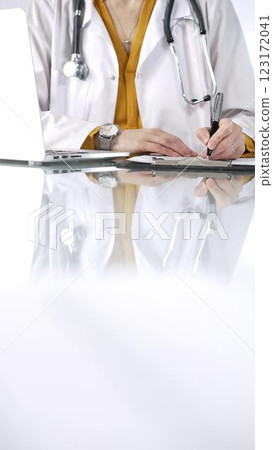 Unknown doctor woman, wearing a stethoscope and yellow blouse under a lab coat, is using a clipboard and taking notes on glass desk in a medical office, close-up vertical view. Medicine concept Unknown doctor woman, wearing a stethoscope and yellow blouse under a lab coat, is using a clipboard and taking notes on glass desk in a medical office, close-up vertical view. Medicine concept 123172041