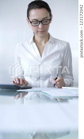 Businesswoman or accountant, wearing white casual blouse and glasses, is working on tax calculations, note taking, and financial reports in the office. Audit and taxes in business Businesswoman or accountant, wearing white casual blouse and glasses, is working on tax calculations, note taking, and financial reports in the office. Audit and taxes in business 123172042
