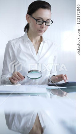 Businesswoman or accountant, wearing white casual shirt and glasses, is using magnifying glass and calculator to analyze financial documents in office setting. Audit and taxes in business Businesswoman or accountant, wearing white casual shirt and glasses, is using magnifying glass and calculator to analyze financial documents in office setting. Audit and taxes in business 123172046
