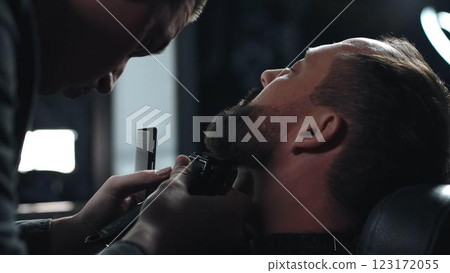 Skilled barber grooming client's beard using electric razor, delivering precise styling within contemporary barbershop setting Skilled barber grooming client's beard using electric razor, delivering precise styling within contemporary barbershop setting 123172055