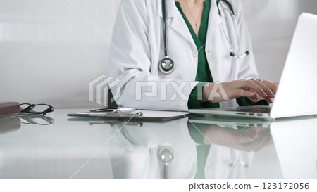 Female doctor using technology in healthcare, close up of hands typing on laptop with stethoscope, glasses, and clipboard on desk 123172056