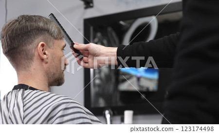 Barber carefully combing hair, client wearing protective cape, preparing for precision styling inside sleek contemporary barbershop setting Barber carefully combing hair, client wearing protective cape, preparing for precision styling inside sleek contemporary barbershop setting 123174498