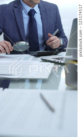 Businessman wearing blue suit is analyzing financial reports with magnifying glass and using calculator in a professional office setting. Low lighting, close-up vertical view. Business people, audit Businessman wearing blue suit is analyzing financial reports with magnifying glass and using calculator in a professional office setting. Low lighting, close-up vertical view. Business people, audit 123174517