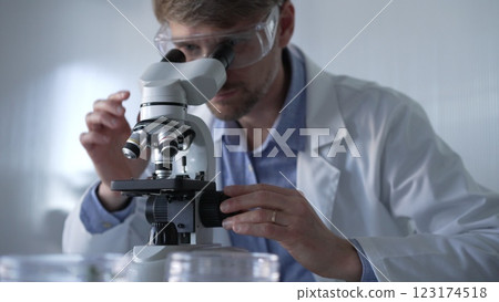 Male scientist wearing lab coat and protective glasses using microscope doing research in modern laboratory setup, portrait view. Low key lighting photo. Science and Medicine concept Male scientist wearing lab coat and protective glasses using microscope doing research in modern laboratory setup, portrait view. Low key lighting photo. Science and Medicine concept 123174518