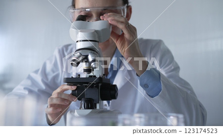 Woman scientist wearing lab coat and protective glasses is using microscope in modern laboratory setup. Low key lighting photo. Science and Medicine 123174523