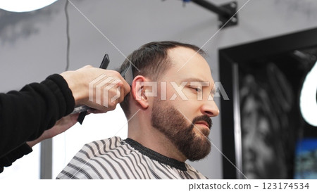 Skilled barber carefully grooming client's beard with electric razor, creating precise, trendy look inside contemporary barbershop setting 123174534