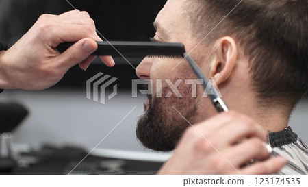 Skilled barber carefully trimming client's beard with precision scissors and sharp comb, working inside contemporary grooming salon 123174535