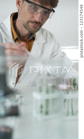 Man scientist wearing a lab coat and protective glasses, is dripping liquid from a pipette into a test tube with a green plant inside, face focused vertical portrait. Science and medicine Man scientist wearing a lab coat and protective glasses, is dripping liquid from a pipette into a test tube with a green plant inside, face focused vertical portrait. Science and medicine 123174548