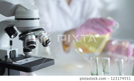 A scientist, wearing a lab coat and pink protective gloves, is pouring a yellow oily liquid from one beaker to another near microscope in laboratory, close up. Medicine and science 123174551