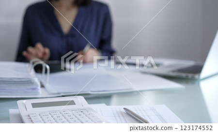 White calculator, pen and documents are lying on accountant's desk. Businesswoman wearing blue dress is working with documents on the background. Business and audit concept White calculator, pen and documents are lying on accountant's desk. Businesswoman wearing blue dress is working with documents on the background. Business and audit concept 123174552