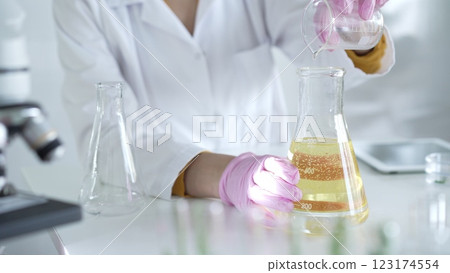 A scientist, wearing a lab coat and pink protective gloves, is pouring a yellow oily liquid from one beaker to another in laboratory, close up. Medicine and science 123174554