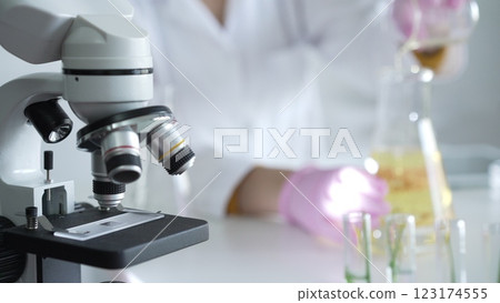 A scientist, wearing a lab coat and pink protective gloves, is pouring a yellow oily liquid from one beaker to another near microscope in laboratory, close up. Medicine and science 123174555