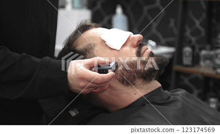 Professional barber grooming client's beard with precision straight razor, delivering classic men's styling in sleek contemporary barbershop setting 123174569