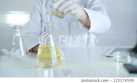 A scientist, wearing a lab coat and white protective gloves, is pouring a yellow oily liquid from one beaker to another in laboratory, close up. Medicine and science concept 123174589