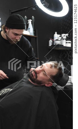 Professional barber carefully shaving client's beard with straight razor, applying gel and using hot towel in contemporary barbershop setting, vertical view Professional barber carefully shaving client's beard with straight razor, applying gel and using hot towel in contemporary barbershop setting, vertical view 123174625