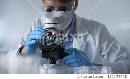 Male scientist researcher wearing blue gloves, protective glasses and medical mask, is using microscope in modern laboratory, portrait view. Low key lighting photo. Science and Medicine 123174638