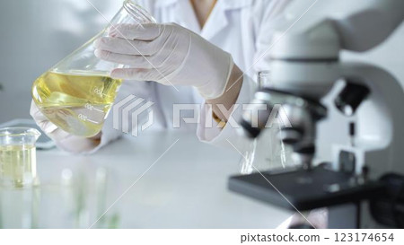 Microscope is near a scientist, wearing a lab coat and white protective gloves, and holding beaker with a yellow oily liquid in laboratory setting, close up. Medicine and science concept 123174654